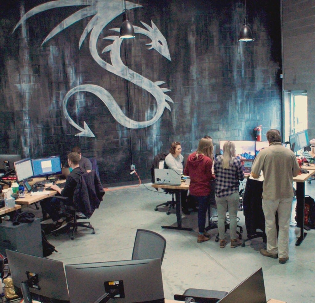 Dragos team members working at the Dragos headquarters in Maryland. There are teams of people standing and sitting at desks. There is a wall with industrial abstract colors and the Dragos logo.