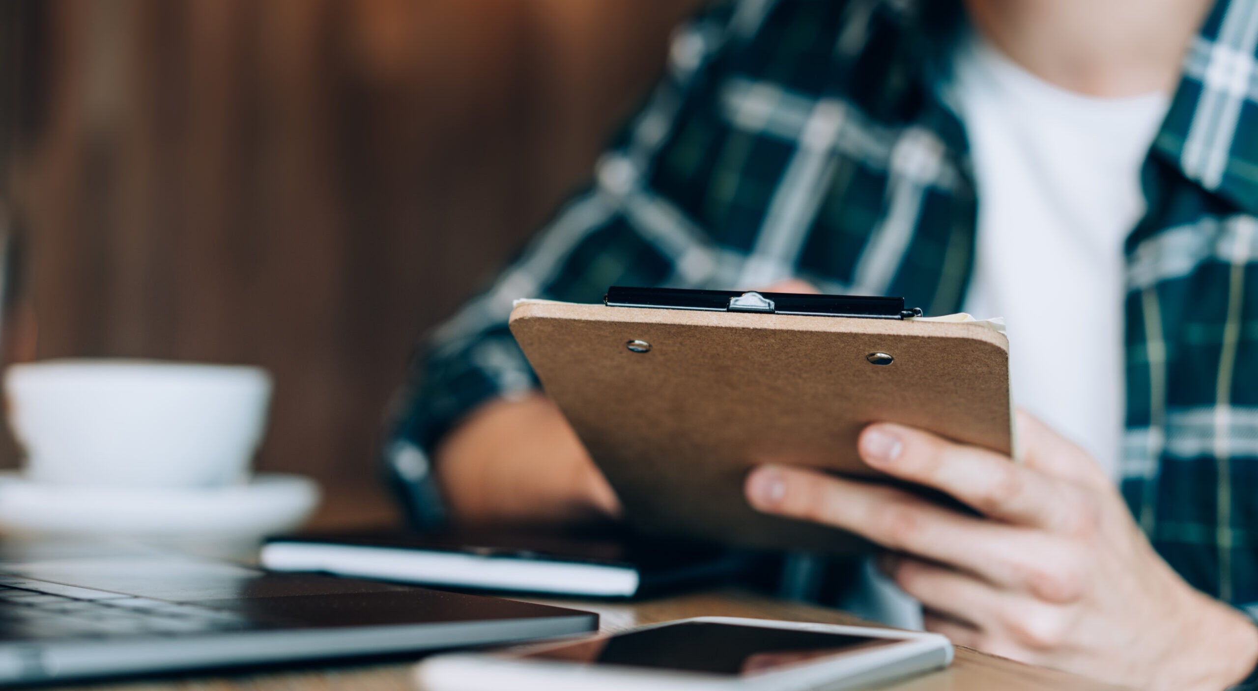 Cropped image of male copywriter making notes of interesting text ideas working remotely in coffee shop, unrecognizable man sitting at table desktop and doing paperwork for analyzing organisation