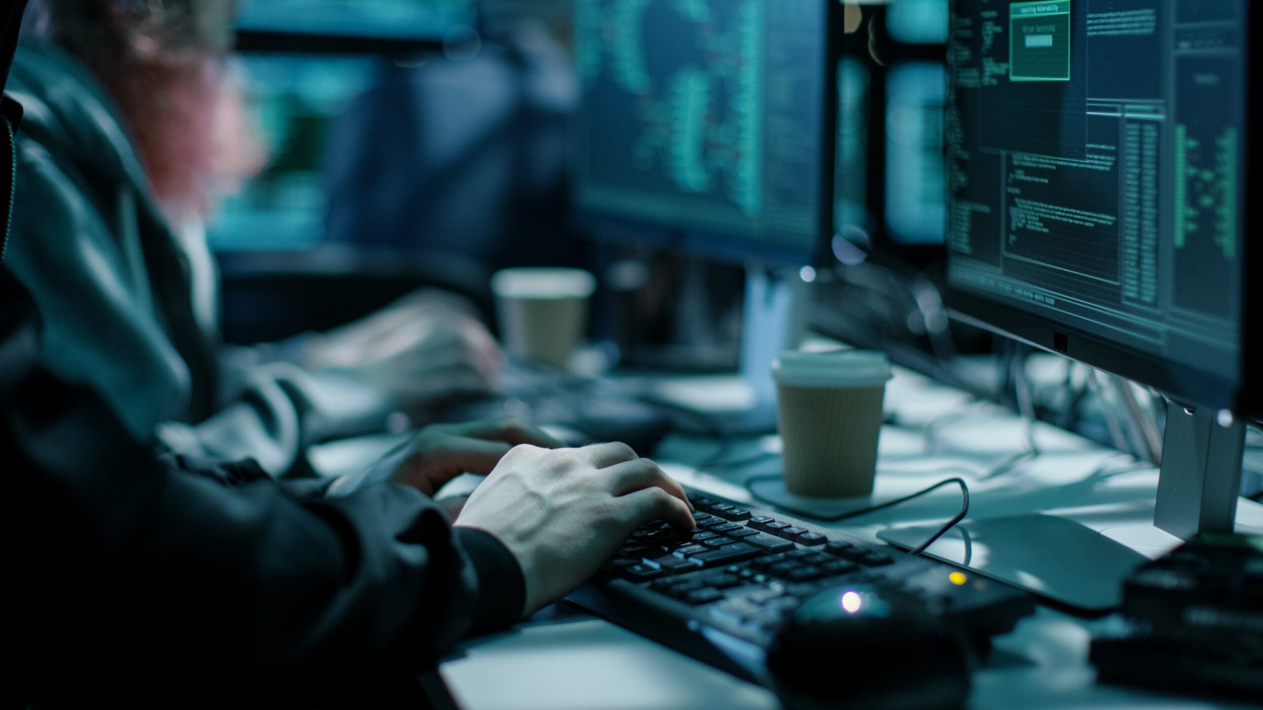 Close-up Shot of Hacker using Keyboard. There is Coffee Cups and Computer Monitors with Various Information.